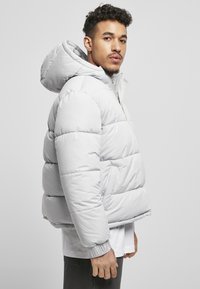 Helle graue Steppjacke mit großer Kapuze, gestepptem Design, gerippten Bündchen und zwei Seitentaschen. Glattes, glänzendes Material.