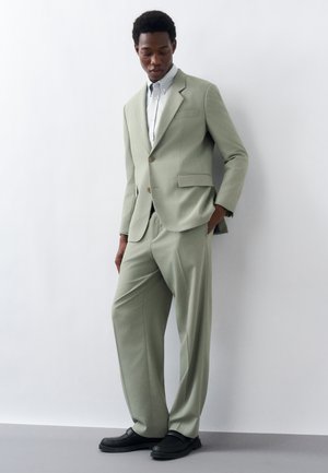 Άνδρας ντυμένος με ανοιχτοπράσινο κοστούμι με φαρδιά παντελόνια, λευκό πουκάμισο με κουμπιά και μαύρα μοκασίνια, στέκεται μπροστά σε λευκό τοίχο.