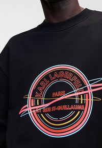 Felpa nera con grafica circolare che presenta "KARL LAGERFELD" in rosso e dettagli in rosa, blu e giallo, su una superficie liscia.