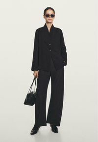 Blazer nero oversize con colletto a scialle, abbinato a pantaloni neri a gamba larga. Gli accessori includono occhiali da sole scuri e una piccola borsa nera.