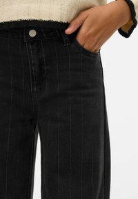 Crne traperice od trapera s bijelim vertikalnim prugama, visokim strukom i prednjim džepovima. Struk ukrašava srebrni gumb.