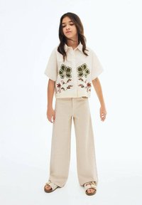 Camicia beige a maniche corte con ricami floreali verdi e chiusura a bottoni, abbinata a pantaloni beige a gamba larga e sandali casual.