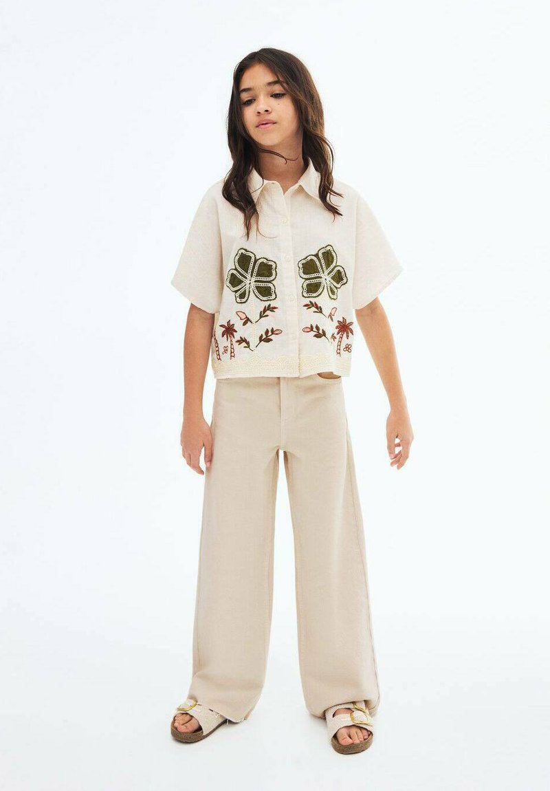 Camicia beige a maniche corte con ricami floreali verdi e chiusura a bottoni, abbinata a pantaloni beige a gamba larga e sandali casual.