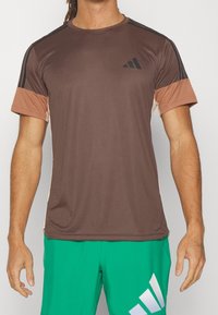 Camiseta atlética marrón de manga corta, con logotipo negro y acentos crema. Tela suave con rayas laterales negras y diseño de cuello redondo.