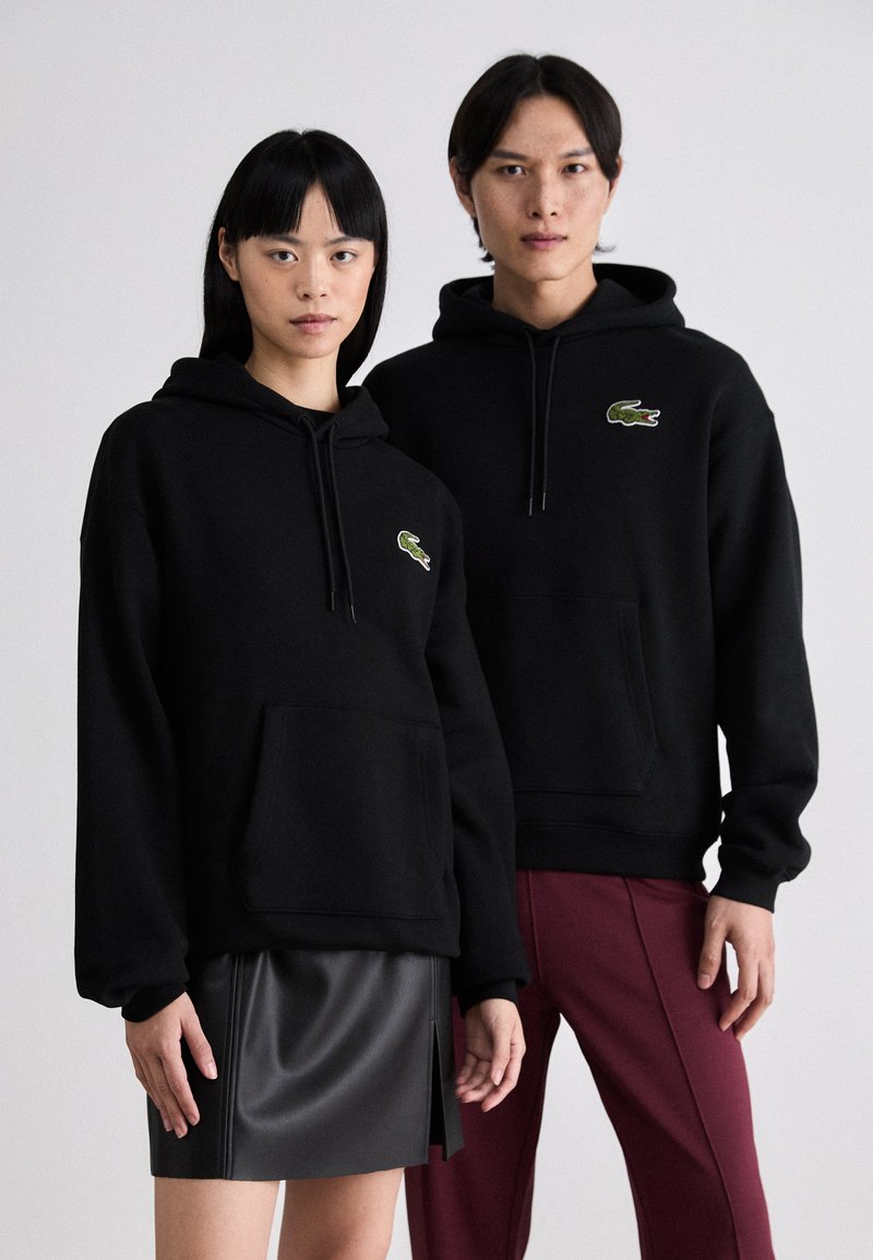 Sudaderas con capucha negras y bolsillos tipo canguro, con un logo verde de Lacoste. Estilizadas con una falda de cuero negra y pantalones burdeos.