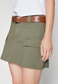 Jupe cargo vert olive avec poches à rabat, ourlet droit et ceinture en cuir brun avec boucle rectangulaire en argent. Tissu en coton lisse.