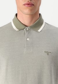 Polo verde oliva com um tecido texturizado, com acabamento branco no colarinho e dois botões. Apresenta um pequeno logótipo da marca bordado no peito.