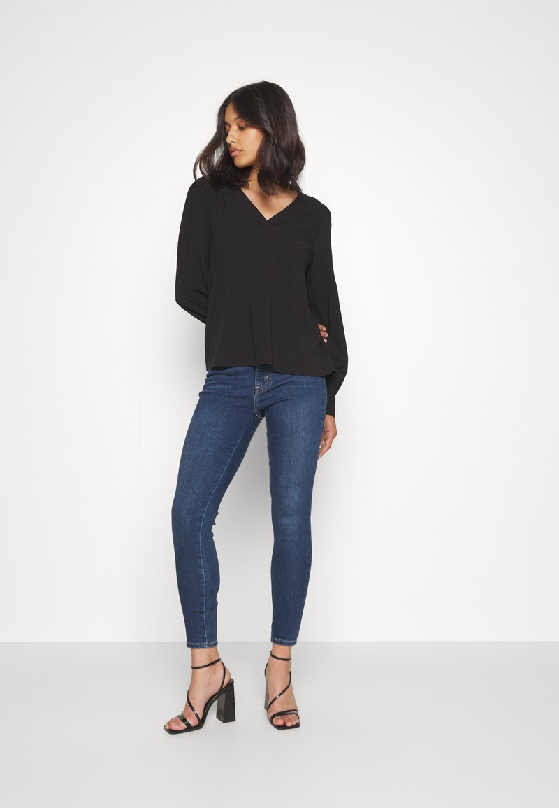 Blouse noire à col en V avec des manches longues, associée à un jean slim bleu foncé et des talons noirs à lanières. Tissu lisse, design ajusté.
