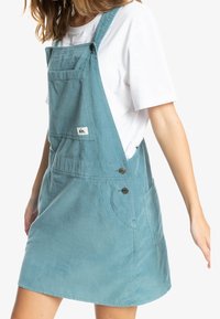 Svetle modré manšestrové šaty na ramínka s nastavitelnými popruhy, přední kapsou a dvěma bočními knoflíky. Hladká textura s ležérním designem.