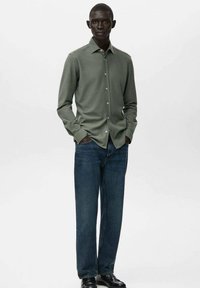 Camisa de manga larga color verde oliva con cuello abotonado, combinada con jeans azules y zapatos negros. Tela suave, diseño informal, forma ajustada.