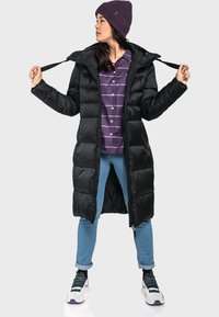 Schwarze Pufferjacke mit Kapuze, mit einem Reißverschluss vorne und elastischen Bündchen. Getragen über einem lila gestreiften Hemd und hellblauen Jeans.