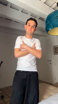 Νεαρός με σγουρά μαλλιά που φοράει λευκό μπλουζάκι και σκούρο παντελόνι στέκεται μέσα σε εσωτερικό χώρο με σταυρωμένα χέρια, δίπλα σε μπλε κρεμαστό φωτιστικό.