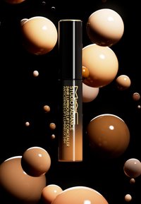 MAC Studio Radiance 24HR Luminous Lift Concealer in einem eleganten schwarzen Tube, umgeben von verschiedenen runden beigen Blasen, vor einem schwarzen Hintergrund.