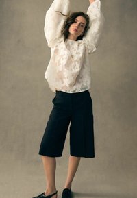 Vit blus med blomstermönster och puffärmar, kombinerad med svarta knälånga shorts. Modellen bär svarta slip-on skor.