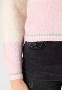 Suéter a rayas en rosa claro y crema con una textura suave, que presenta un dobladillo y puños de canalé con rayas de acento en gris y blanco. Combinado con pantalones de mezclilla negros.