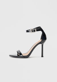 Sandalia de tacón alto en piel sintética negra con correa en el tobillo. Presenta un diseño minimalista, punta abierta y una hebilla con detalles de pedrería.