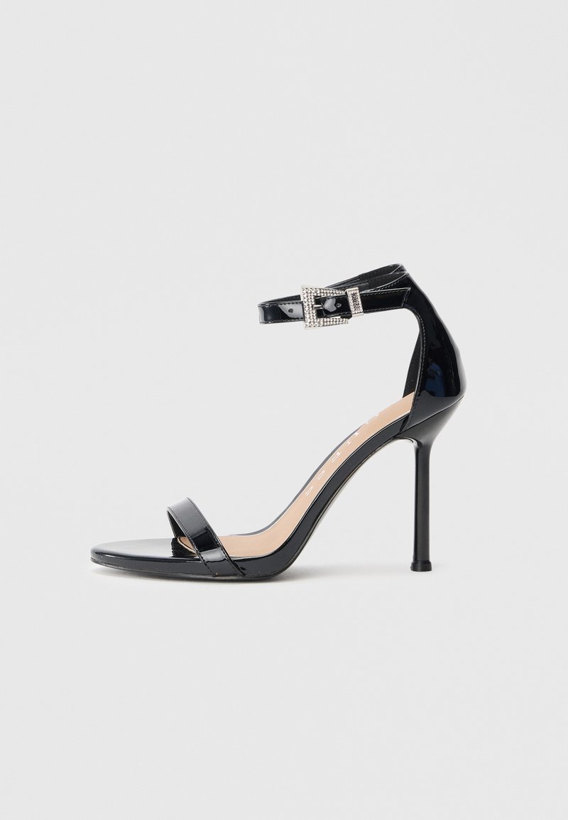 Sandalia de tacón alto en piel sintética negra con correa en el tobillo. Presenta un diseño minimalista, punta abierta y una hebilla con detalles de pedrería.
