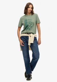 Groen katoenen T-shirt met een grafische opdruk, gecombineerd met rechte blauwe denim jeans. Licht truitje gestrikt rond de taille en zwarte schoenen.