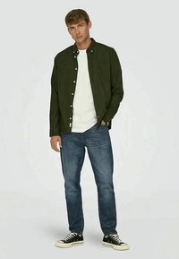 Camisa verde escura de botões sobre uma t-shirt branca, combinada com jeans de ganga azuis e ténis pretos. Design simples com um corte casual.