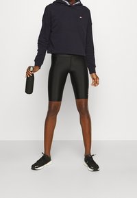 Sudadera corta negra con detalle del logo, pantalones cortos negros elásticos y zapatillas deportivas negras. Sostiene una botella de agua negra.