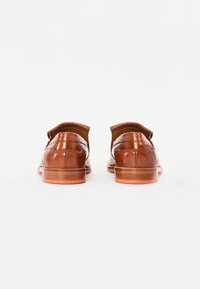 Zapatos de cuero marrón con una textura suave, con la parte trasera redondeada y suelas de goma naranja. El diseño está acentuado con costuras decorativas.