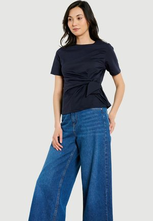 Vrouw draagt een marineblauw kort mouwen top met een zijstrik en wijde blauwe spijkerbroek, staand tegen een lichtgrijze achtergrond.