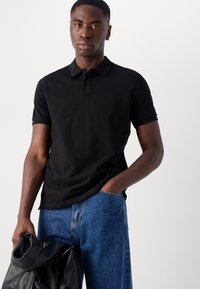 Schwarzes Poloshirt mit Kragen, kurzen Ärmeln und einem dezenten Logo. Kombiniert mit blauen Jeans und hält eine schwarze Lederjacke.