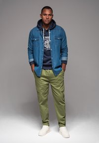 Jeanshemd über einem dunkelblauen Hoodie, kombiniert mit olivgrünen Cargo-Hosen und weißen Sneakers. Verfügt über zwei Brusttaschen und einen Kordelzugbund.