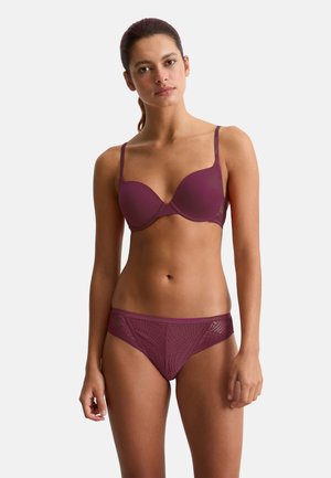 Frau mit braunen Haaren im Pferdeschwanz, die einen passenden lila BH und Spitzenunterhose trägt, vor einem einfarbigen hellen Hintergrund stehend.