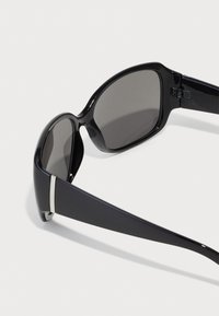 Schwarze Sonnenbrille mit einem eleganten Kunststoffrahmen, dunklen Gläsern und silbernen Akzenten an den Schläfen, die ein geschwungenes Design und eine glatte Textur aufweisen.