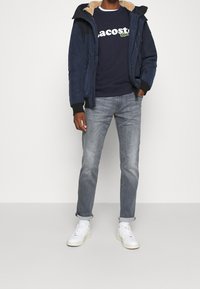 Marinblå parkas med pälsfoder i huvan, marinblå t-shirt med vitt "Lacoste"-logotyp, grå jeans och vita sneakers. Avslappnad lager-outfit.