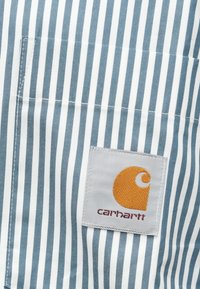 La tela de rayas azul y blanca presenta un bolsillo en el pecho izquierdo y un parche de marca con un logo naranja y el texto "carhartt".