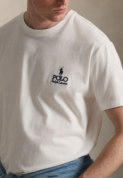 Junge Person trägt ein weißes Polo Ralph Lauren T-Shirt mit gesticktem Logo auf der Brust, kombiniert mit hellblauen Jeans, neutraler Hintergrund.