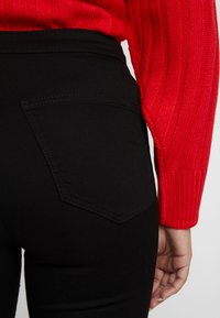 Pantalones negros con un diseño ajustado y un detalle de bolsillo trasero, combinados con un suéter de punto acanalado rojo. Enfóquese en el material elegante y los colores contrastantes.