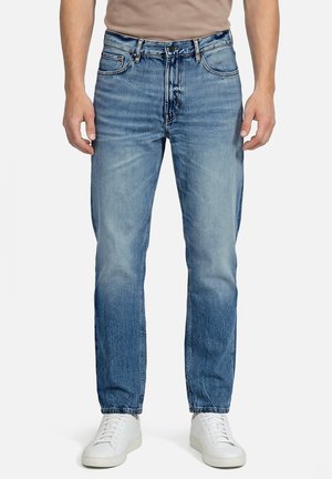 Mann trägt hellblaue ausgewaschene Jeans mit einem beigen Hemd und weißen Sneakers, steht vor einem schlichten weißen Hintergrund.