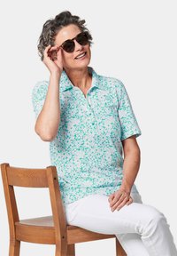 Korte mouwen polo shirt in lichtblauw met een bloemmotief in verschillende tinten teal. Model met zonnebril, zittend op een houten stoel.
