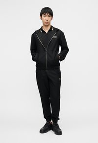 Μαύρο αθλητικό σετ από υλικό με υφή, που περιλαμβάνει φούτερ με φερμουάρ και παντελόνια τζόγκερ με λογότυπο. Υπόδηση: μαύρα αθλητικά παπούτσια.