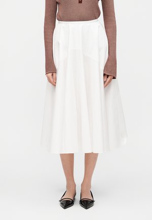 Femme portant une jupe mi-longue blanche en ligne A avec des poches avant, un haut côtelé marron à manches longues et des ballerines Mary Jane noires à bout pointu.