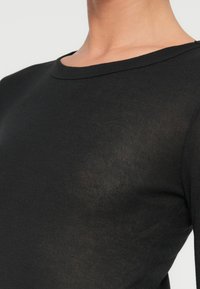 Γυναίκα που φοράει εφαρμοστή μαύρη μπλούζα με μακριά μανίκια και στρογγυλή λαιμόκοψη, που αφήνει εμφανή τον λαιμό και το πάνω μέρος του κορμού.