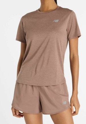 Kobieta w brązowej sportowej koszulce z krótkim rękawem i dopasowanych szortach z logo New Balance, stojąca na jednolitym białym tle.