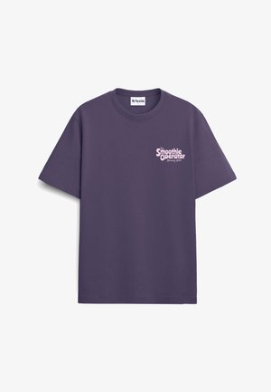 Osoba z kręconymi włosami nosi fioletowy T-shirt z różowym napisem i kreskówkową postacią smoothie na deskorolce na plecach.