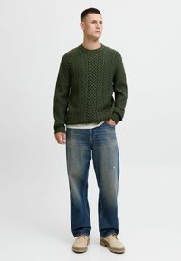 Donkergroen gebreide trui met een textuur kabelontwerp, gecombineerd met lichtblauwe versleten spijkerbroek en beige schoenen. Casual pasvorm en ontspannen stijl.