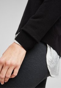 Gros plan d'une personne portant un pull noir sur une chemise blanche, reposant sa main sur un pantalon foncé texturé.