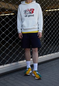Felpa grigia con logo "New Balance", maglietta gialla, pantaloni corti blu navy, calze bianche e scarpe multicolori con dettagli verdi e gialli.