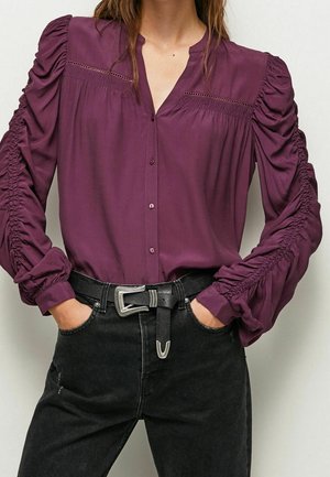 Kvinde iført en dyb lilla bluse med lange ærmer med rynkede ærmer og sorte jeans med sort bælte med sølvspænde.