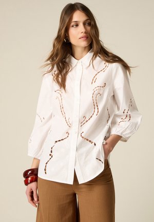 Kobieta w białej bluzce na guziki z wyciętymi kwiatowymi wzorami i luźnymi rękawami, zestawiona z brązowymi spodniami o szerokich nogawkach i czerwonymi bransoletkami.