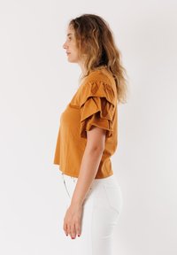 T-shirt corto marrone con maniche a volant, realizzato in morbido tessuto. Abbinato a pantaloni bianchi, che mostrano una silhouette pulita e semplice.