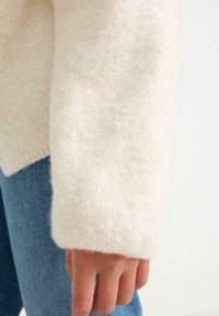 Cremefarvet sweater lavet af blødt, struktureret stof med brede, ribbede manchetter; parret med lyseblå jeans. Nærbillede af ærmet og hånden.