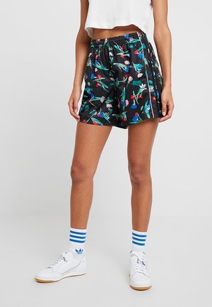 Frau trägt schwarze Shorts mit Blumenmuster, weißes kurzes Top, weiße Sneaker und weiße Socken mit blauen Streifen, steht vor einem schlichten Hintergrund.