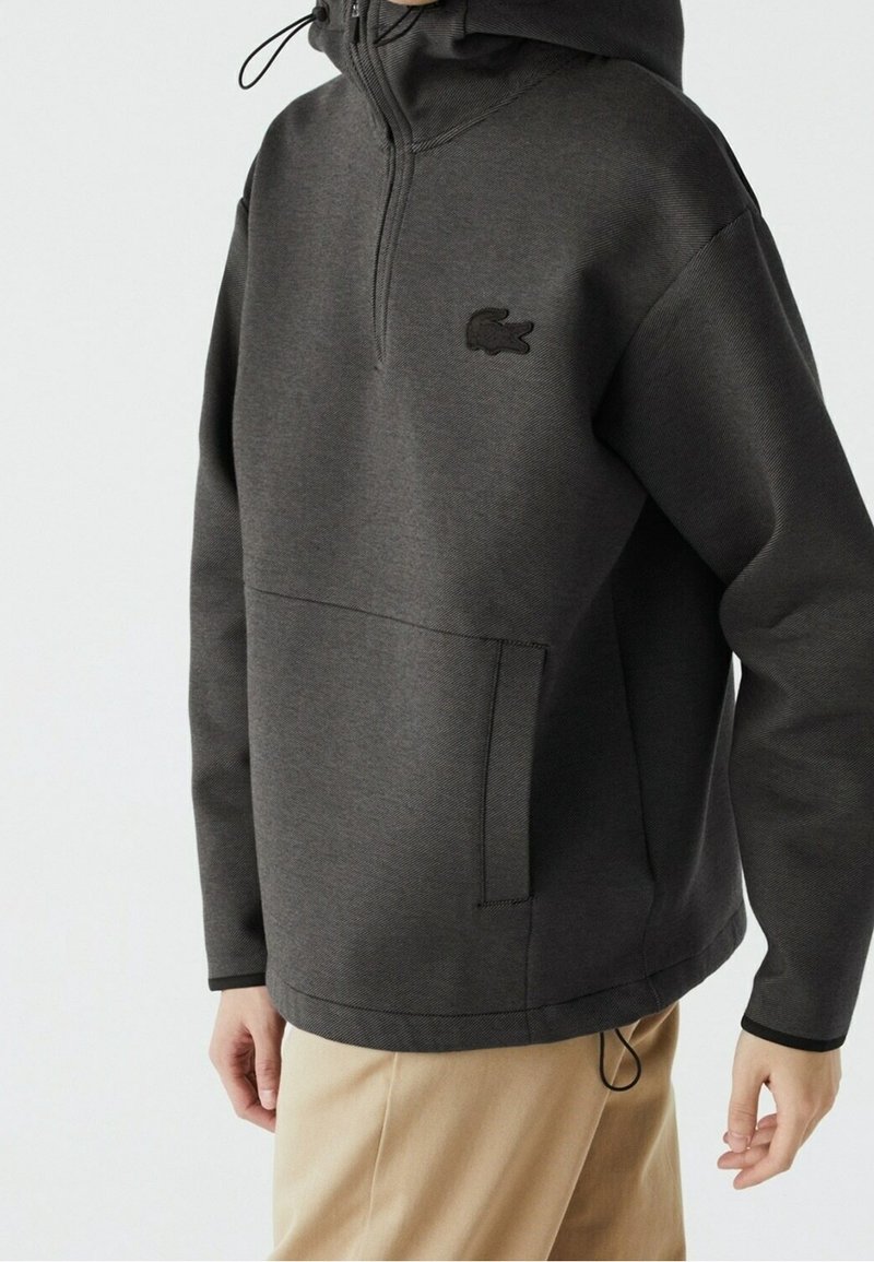 Sweat à capuche gris foncé de poids moyen avec une poche avant, un col à fermeture éclair sur un quart, et un petit logo noir sur la poitrine. Capuche ajustable avec cordon de serrage.